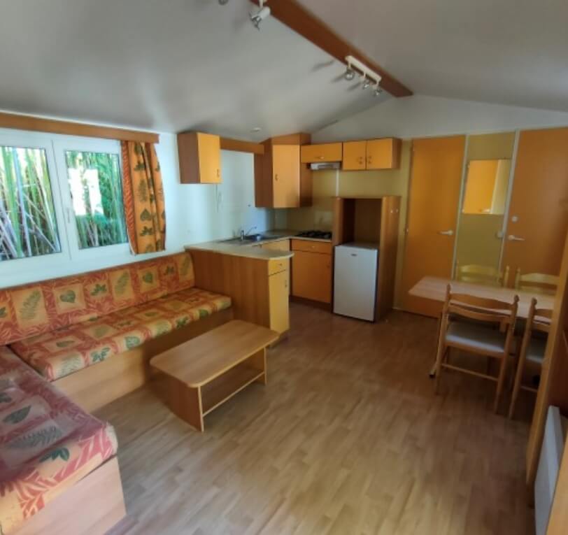 View of the kitchen, dining area and living area in the ‘Le Truffier’, ‘Le Cornouiller’, ‘Le Noisetier’ and ‘L’Olivier’ mobile homes available for hire at Le Bel Air campsite in the Lot, featuring a semi-covered wooden terrace